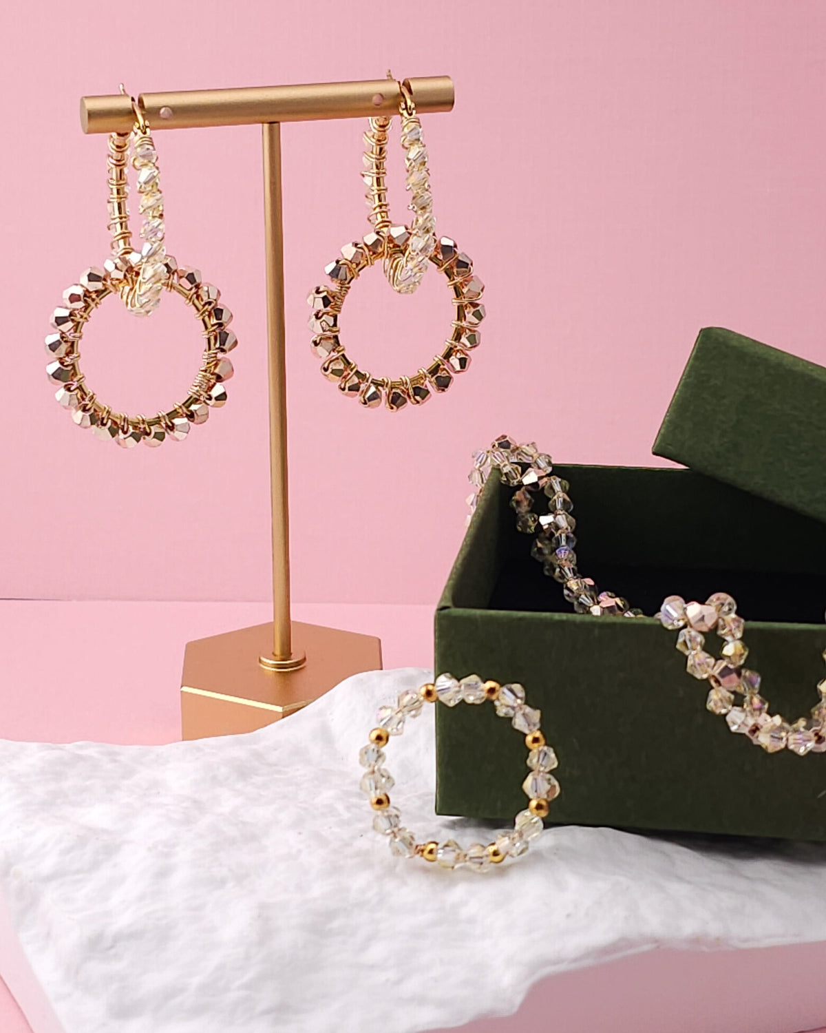 Gold-tone beaded hoop earrings displayed on a gold stand, with a matching beaded bracelet and a dark green jewelry box on a pink background.