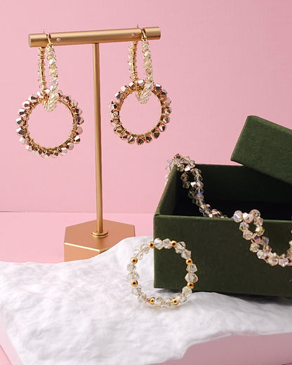 Gold-tone beaded hoop earrings displayed on a gold stand, with a matching beaded bracelet and a dark green jewelry box on a pink background.