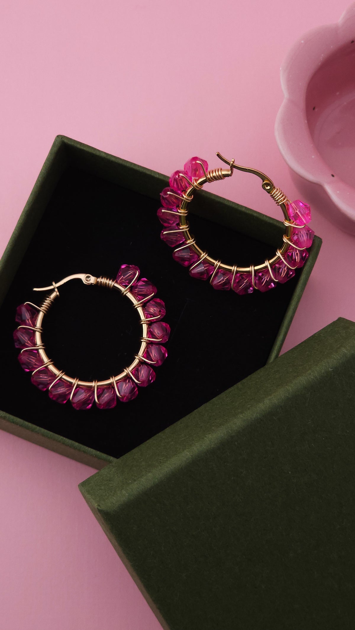 Gold hoop earrings with a textured pink crystal border, presented in an open dark green jewelry box on a pink background.